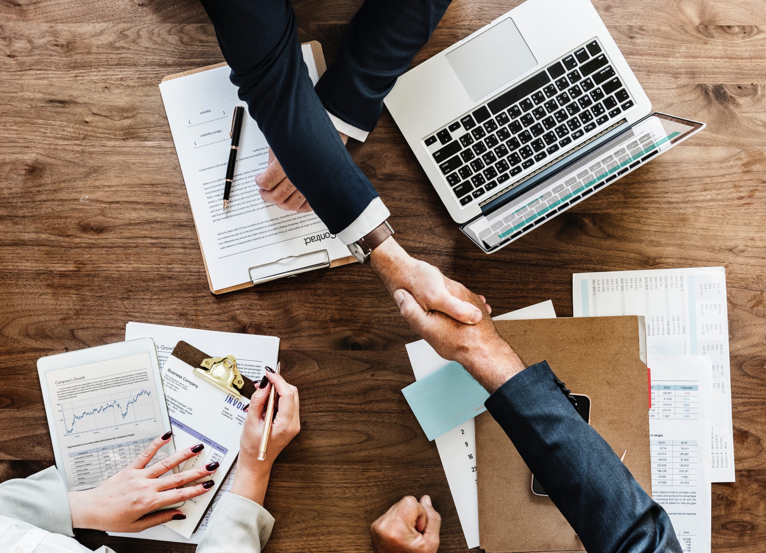 2 business men shaking hands round the table . laptop, pen and papers placed on wooden brown table